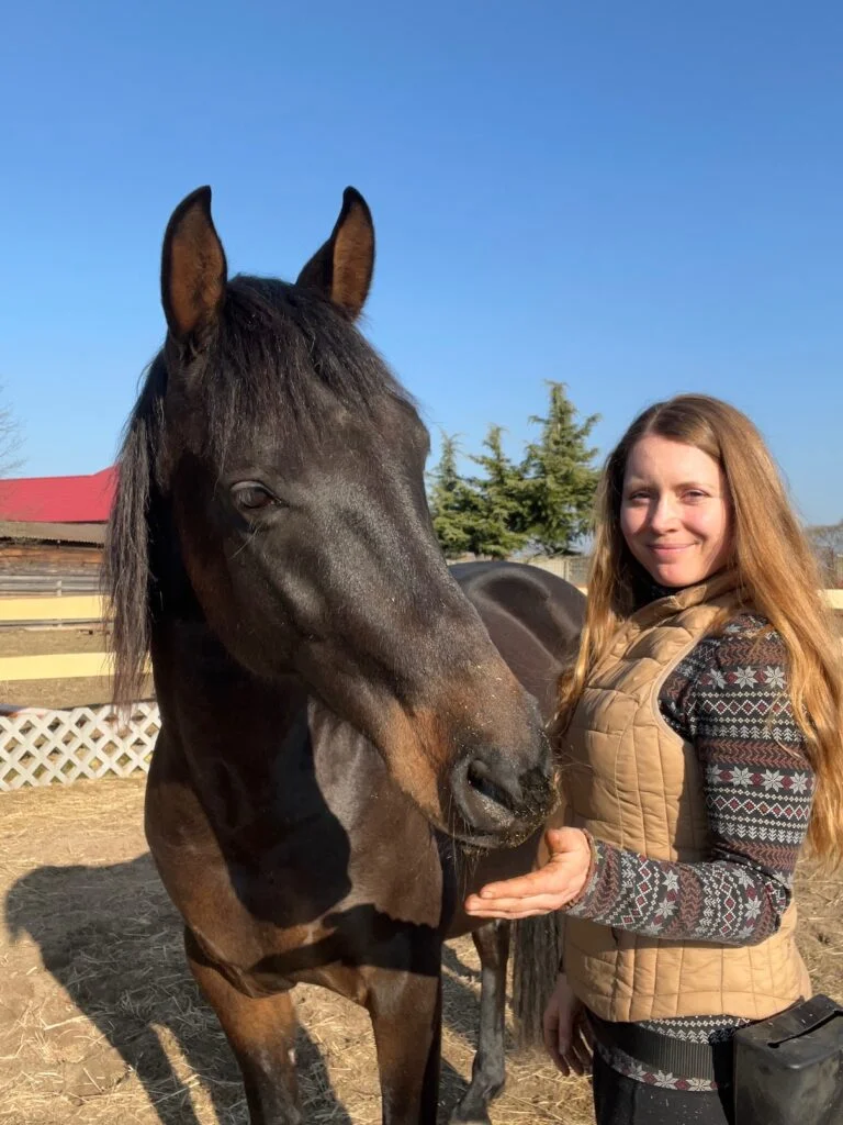 Žena hladí hnědého koně na slunečním dvorku s dřevěným plotem a stromy v pozadí.